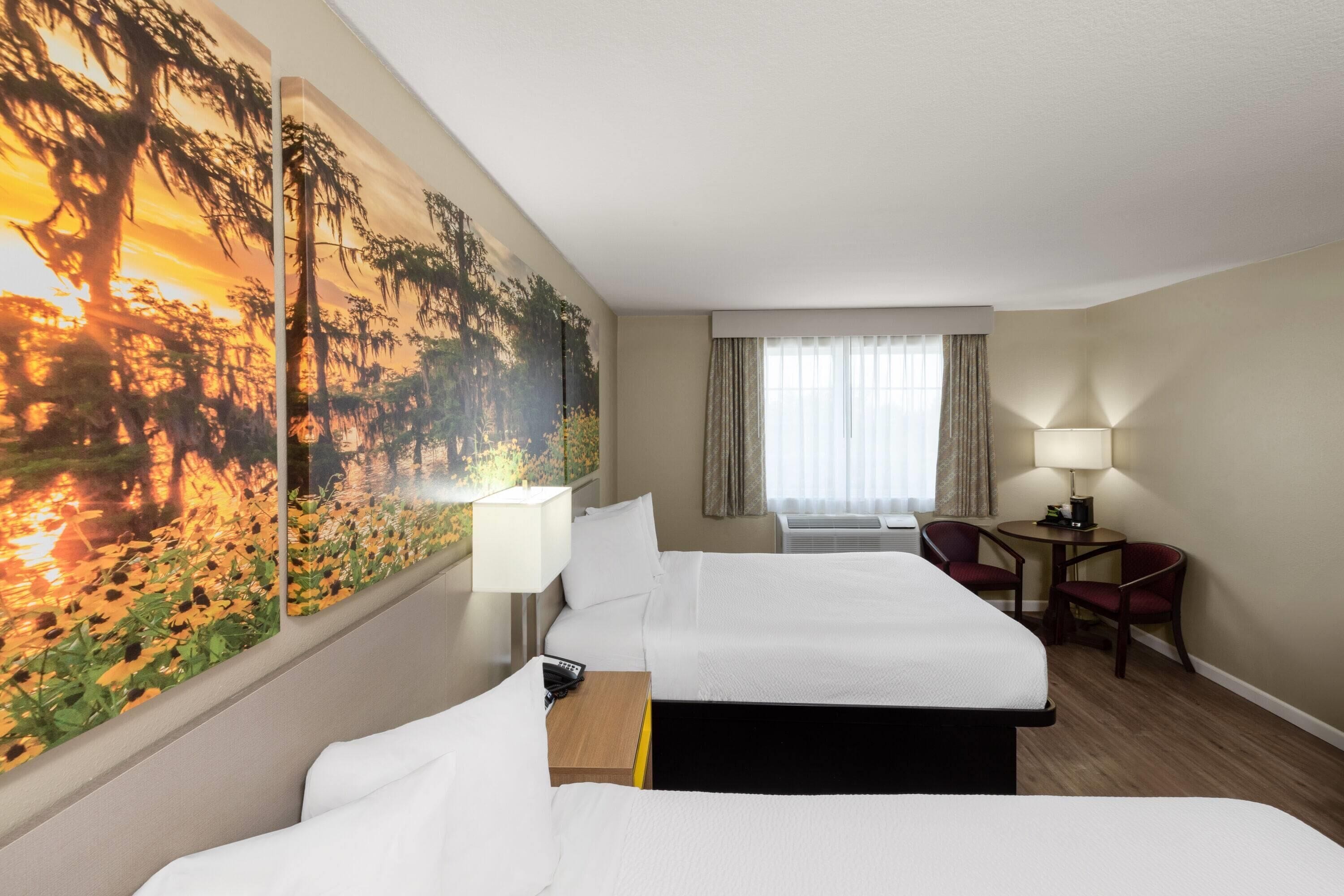 A hotel room with two beds, a desk with a lamp, a chair, a large painting on the wall, and a window with curtains.