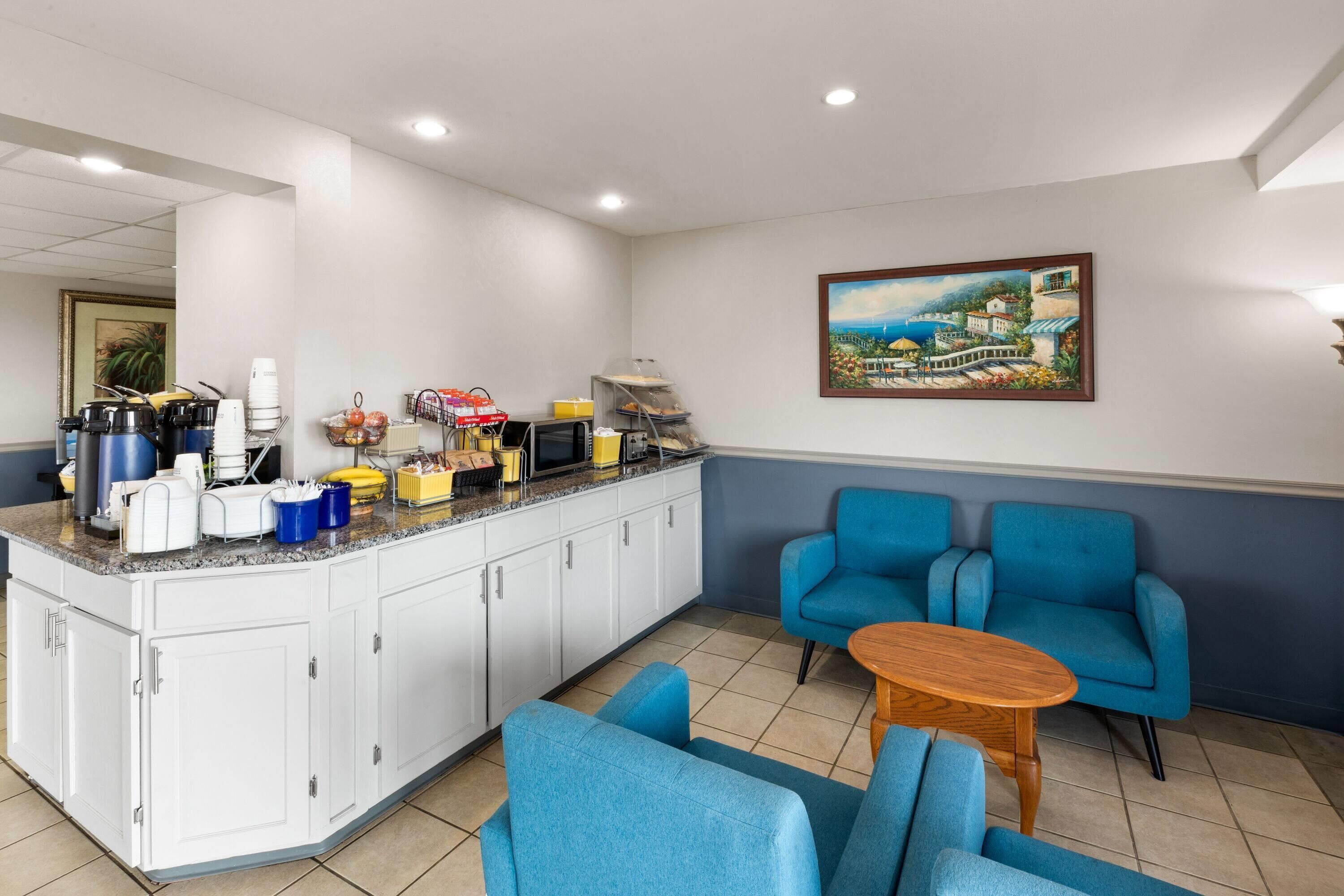 A kitchen with white cabinets, a microwave, and various appliances. There are blue chairs and a small wooden table. A painting of a coastal scene is on the wall.