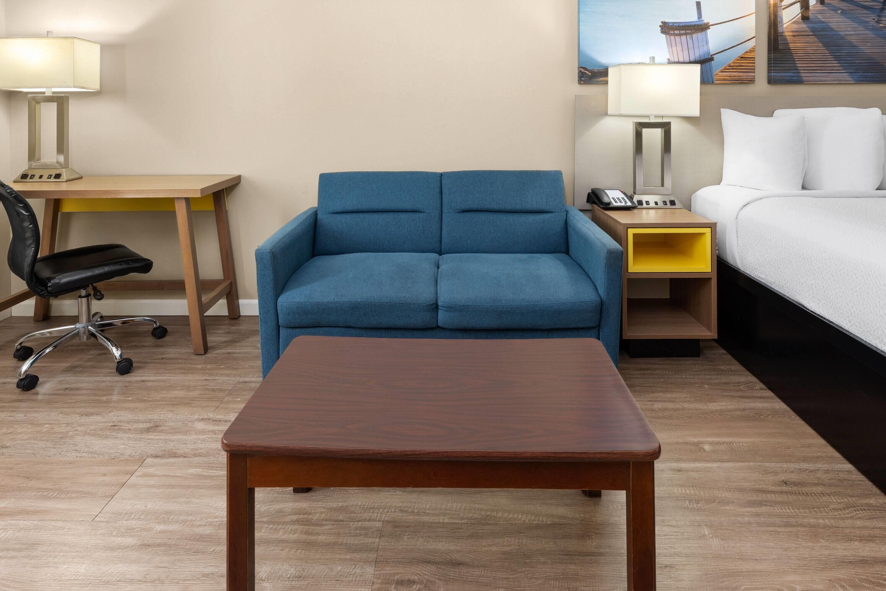 A hotel room with a bed, a blue sofa, a wooden table, a desk with a lamp, and a chair.