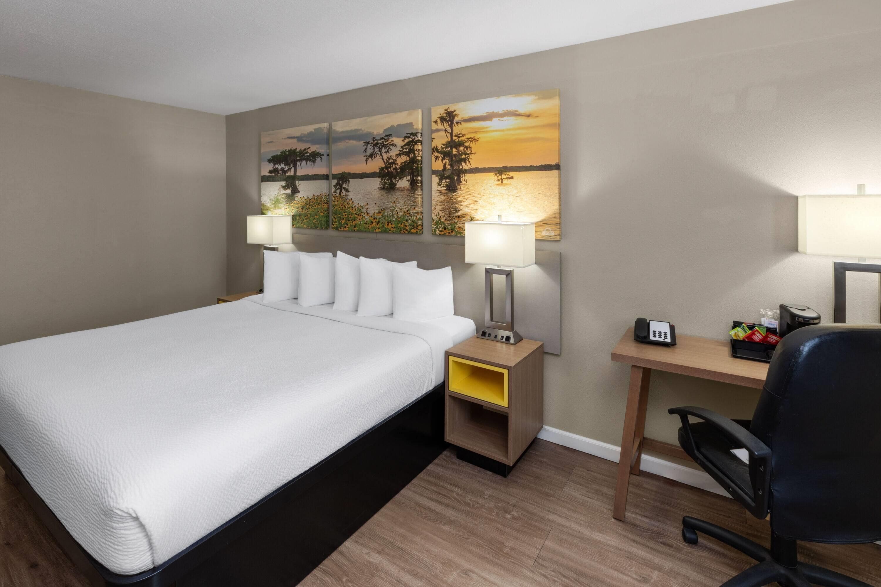A hotel room with a large bed, two bedside tables with lamps, a desk with a chair, and a wall with three paintings.