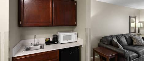 Una cocina compacta con un lavamanos, microondas y refrigerador negro. Junto a la cocina, un sofá de cuero oscuro con un reposapiés a juego.