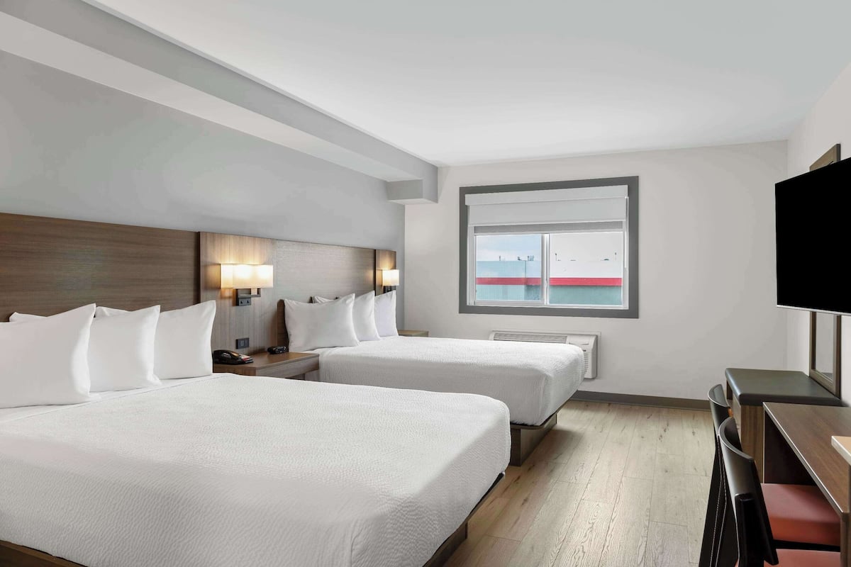 Ein Hotelzimmer mit zwei Betten, einem Schreibtisch, einem Fernseher und einem Fenster mit Blick auf ein Gebäude.