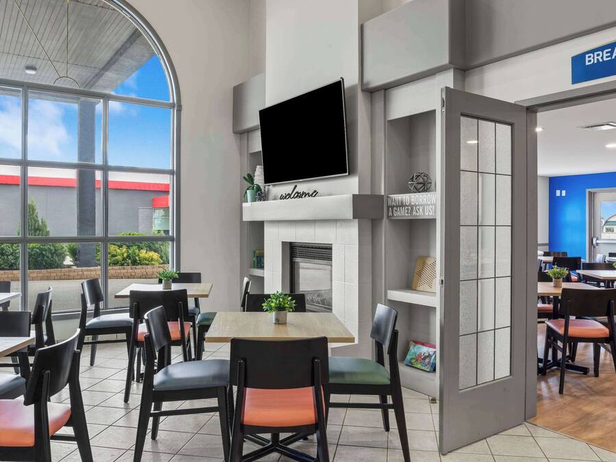 A modern dining area with tables and chairs, a fireplace, and a large window offering a view of the outside.