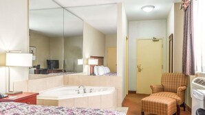 Ein Hotelzimmer mit einem großen Bett, einer Sitzecke mit einem Sessel, einem großen Spiegel, einer Badewanne und einer Tür zu einem weiteren Raum.