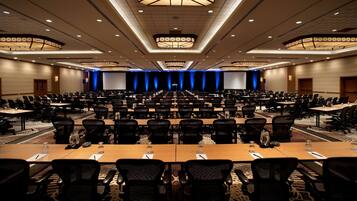 A large conference room with rows of tables and chairs, a stage with a blue curtain backdrop, and a modern chandelier.