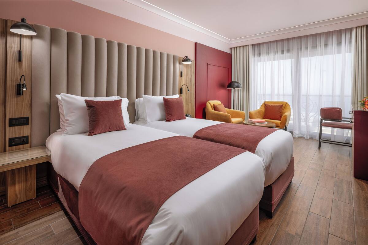 A hotel room with two beds, a desk, a chair, and a large window with curtains.