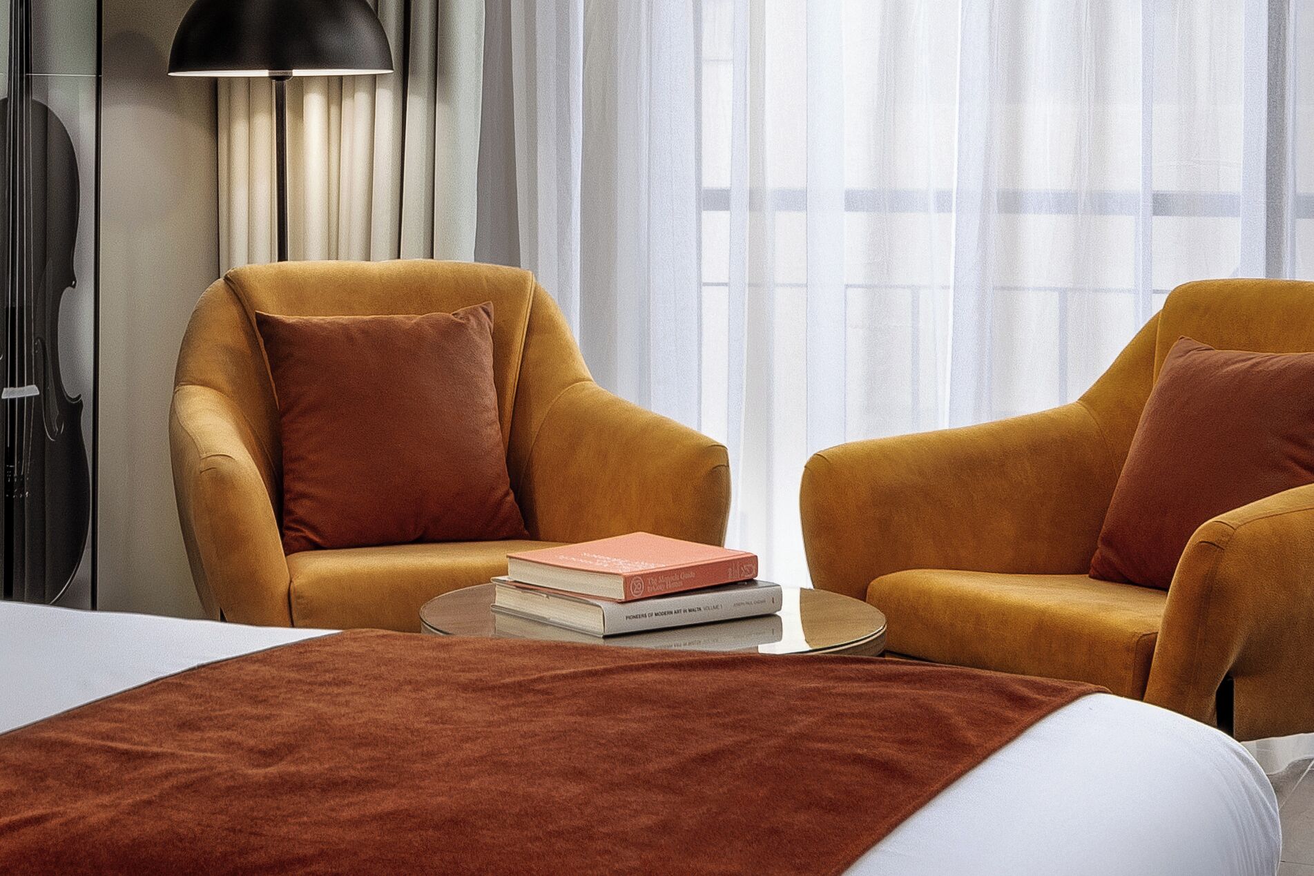 A hotel room with two armchairs, a bed, a bedside table with books, a lamp, and a window with sheer curtains.