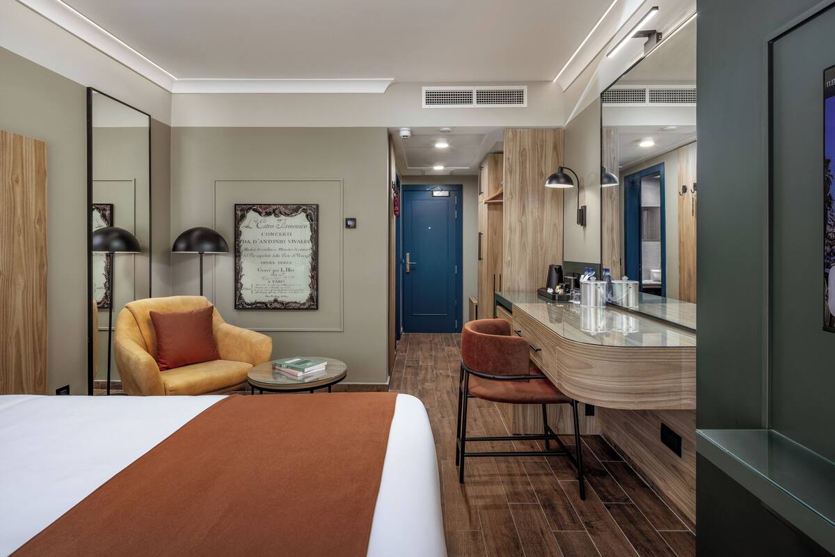 A hotel room with a bed, a chair, a small table, a mirror, a TV, and a door leading to another room.