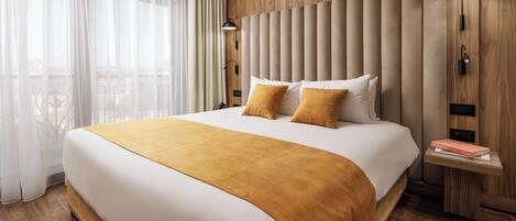 A modern hotel room with a large bed, wooden paneling, a nightstand with a lamp, and a view of the outdoors.