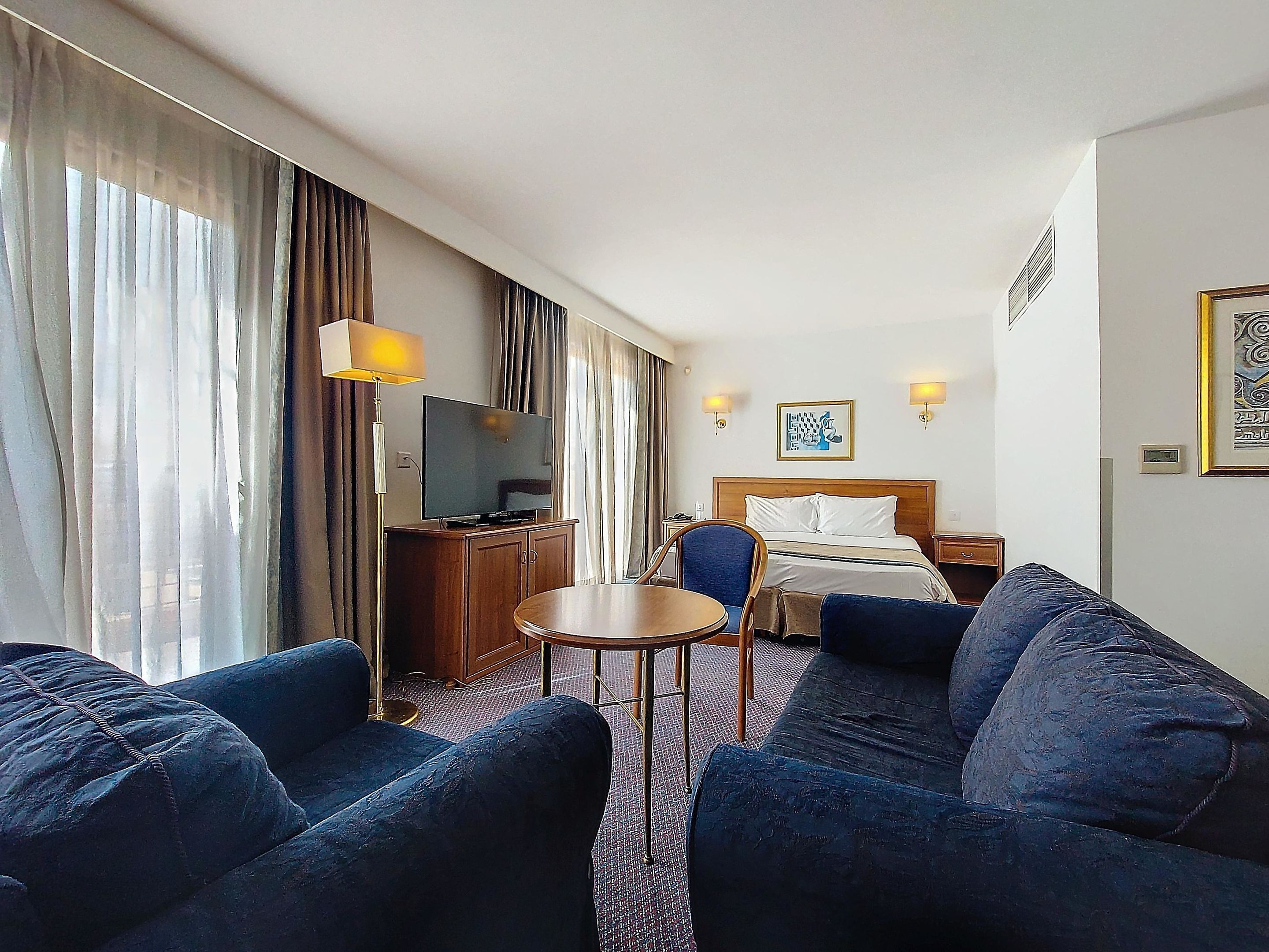 Habitación de hotel con cama, escritorio, televisor, dos sofás, una mesita y cortinas.