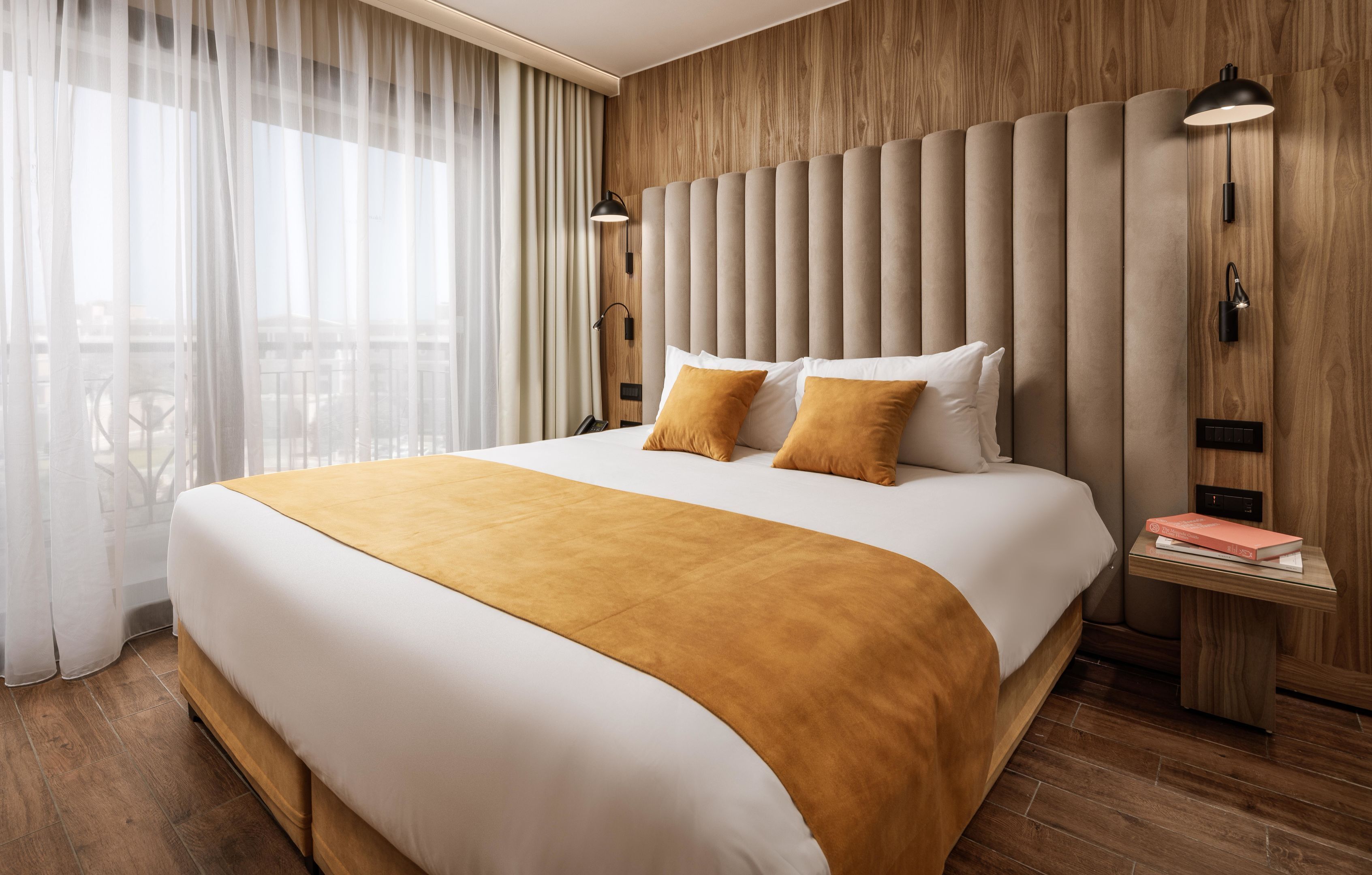 Een moderne hotelkamer met een groot bed, houten lambrisering, een nachtkastje met een lamp en uitzicht door dunne gordijnen.