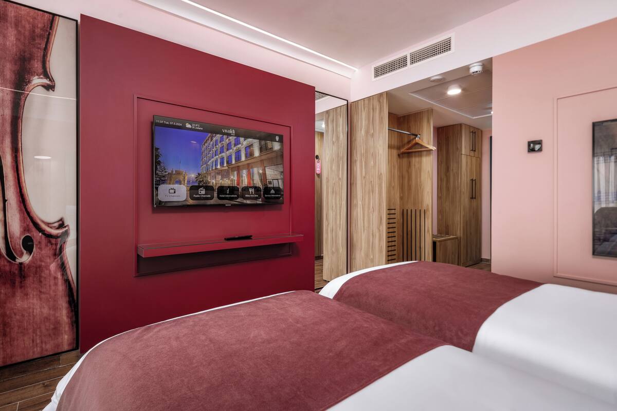 A hotel room with a TV mounted on a red wall, wooden wardrobe, and a bed with a red and white duvet.