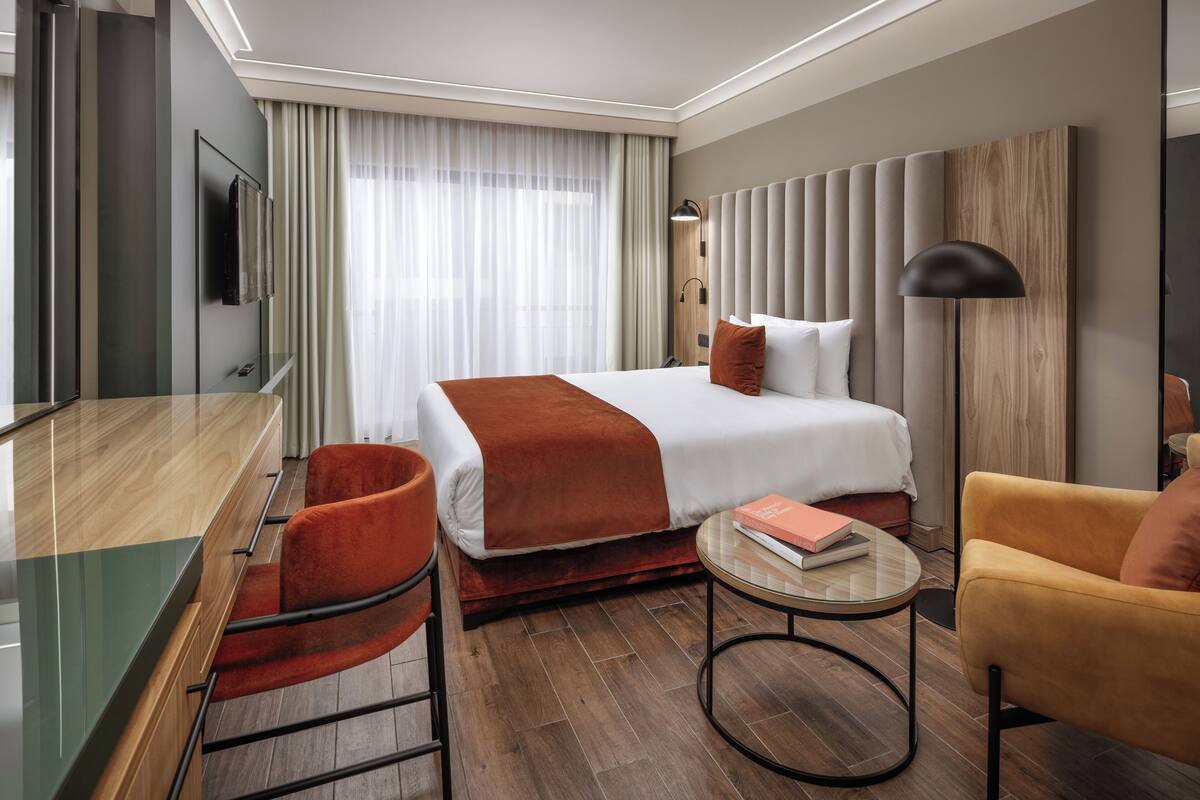 A modern hotel room with a large bed, a desk, a chair, and a round table.