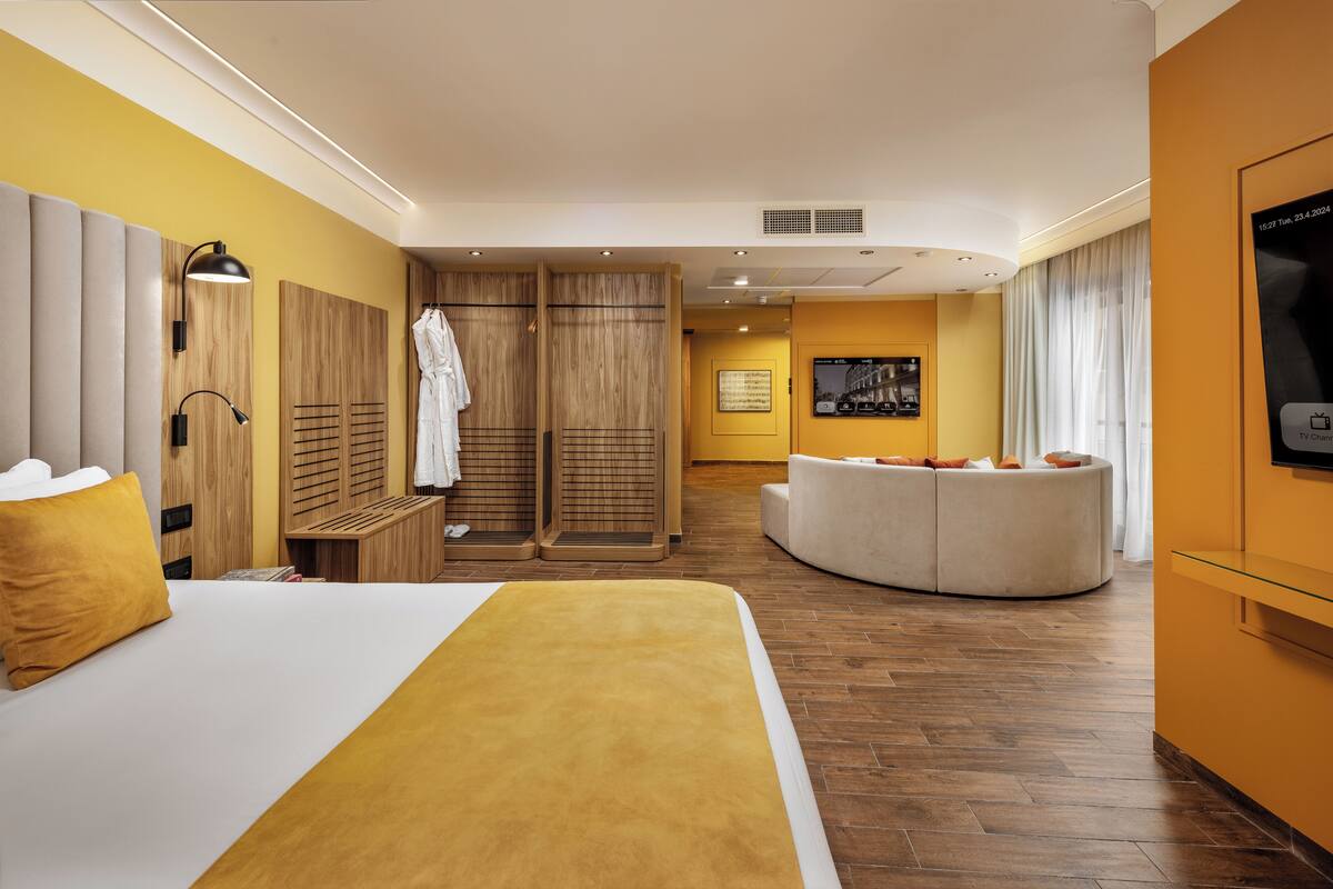 A hotel room with a large bed, a seating area with a sofa, wooden wardrobes, a TV, and a wall-mounted TV.
