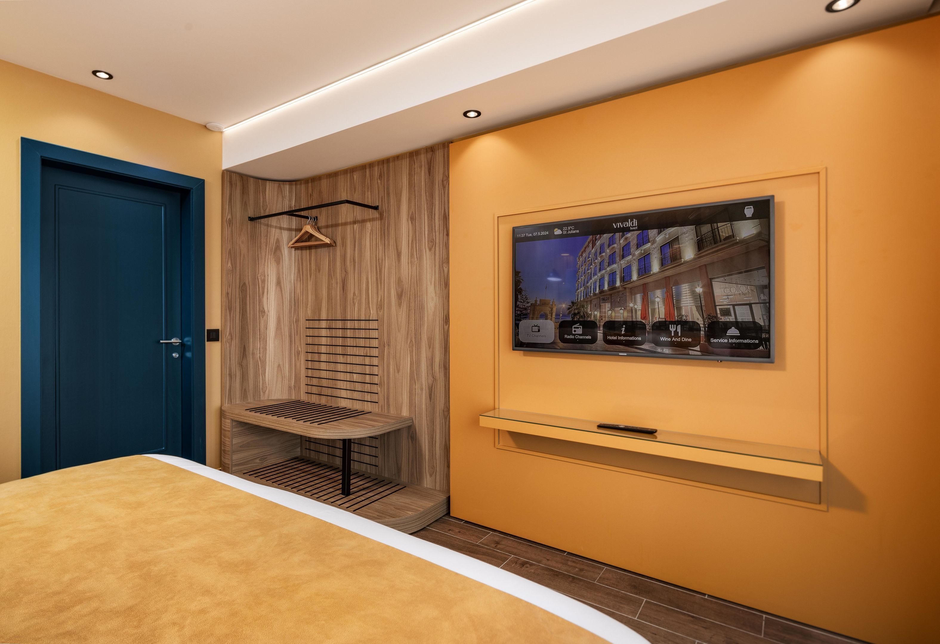 A hotel room with a blue door, a wooden panel wall, a mounted TV, and a small shelf.