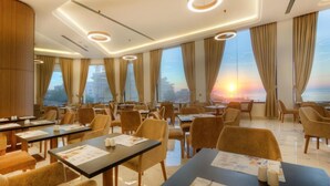 A hotel dining area with tables and chairs, large windows with curtains, and a sunset view.