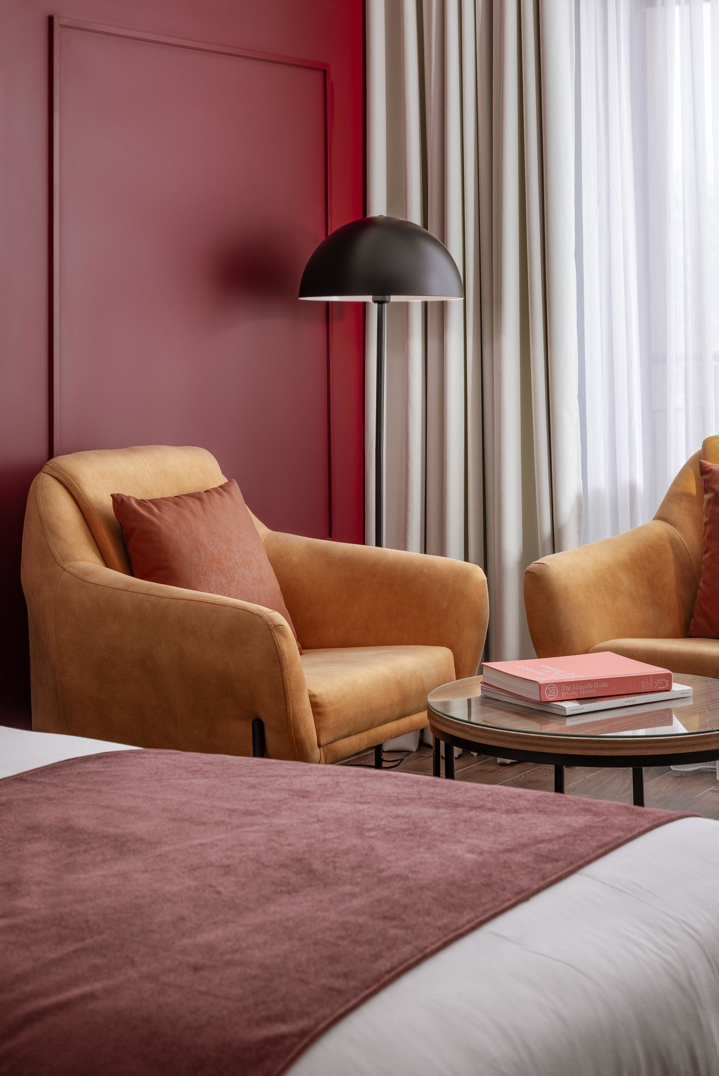 A hotel room with a bed, two armchairs, a round coffee table, a floor lamp, and a large window with curtains.