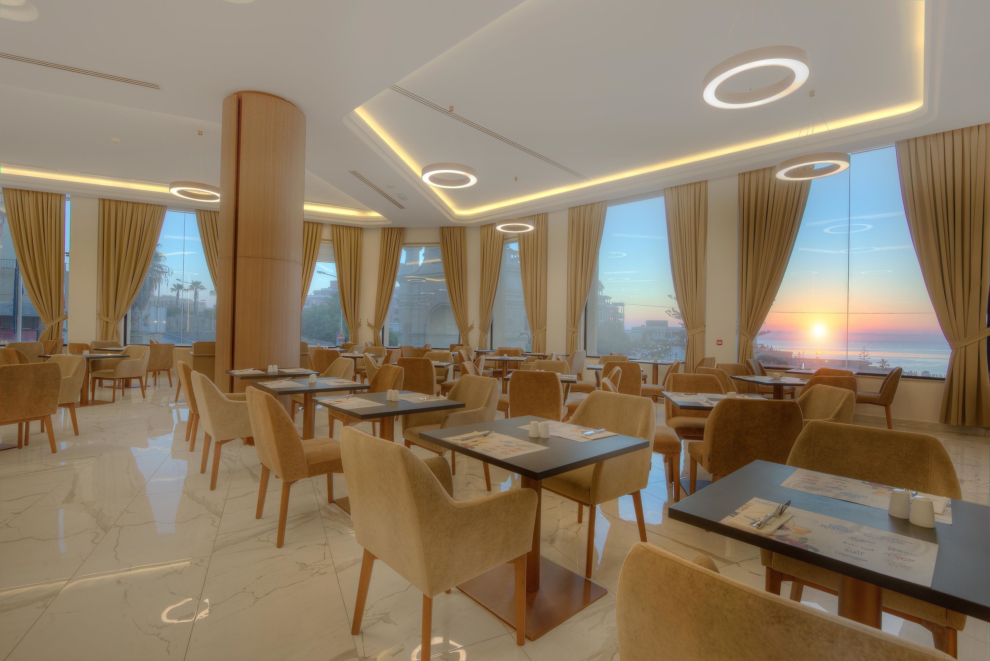 A hotel dining area with tables, chairs, and large windows offering a sunset view.