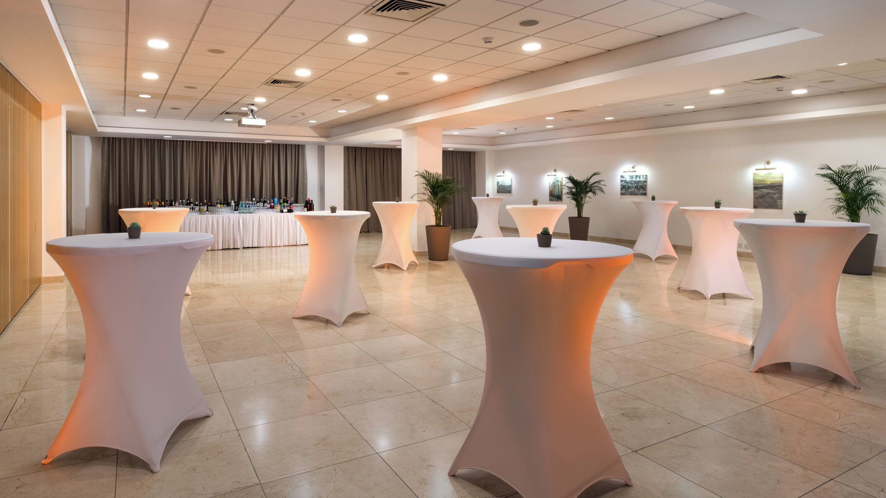 A spacious room with multiple round tables, a bar area with bottles, and potted plants.