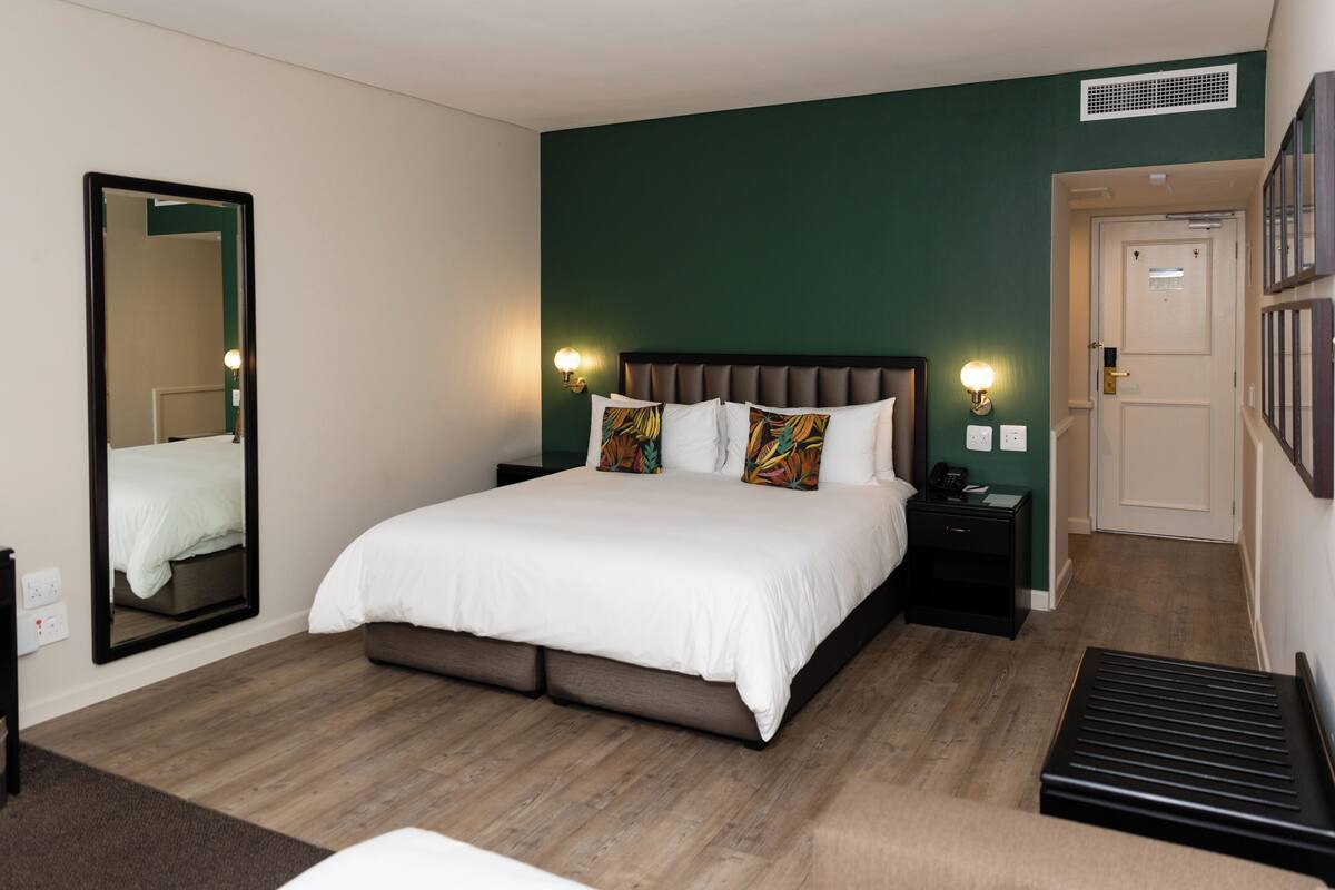 A hotel room with a large bed, a bedside table, a nightstand, a mirror, and a door leading to another room.
