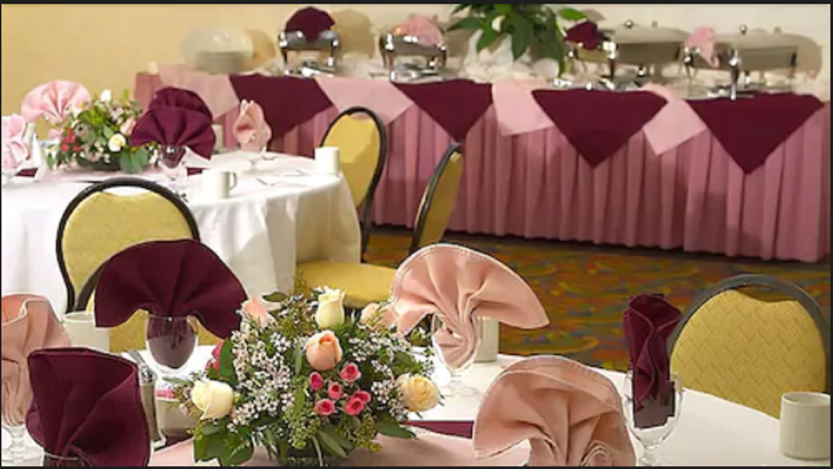 A banquet hall with round tables covered in white tablecloths and pink table runners, chairs with yellow and pink upholstery, and floral centerpieces.