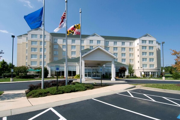A hotel with a central entrance, multiple flags, and a parking lot. - Hilton Garden Inn Owings Mills (Owings Mills)