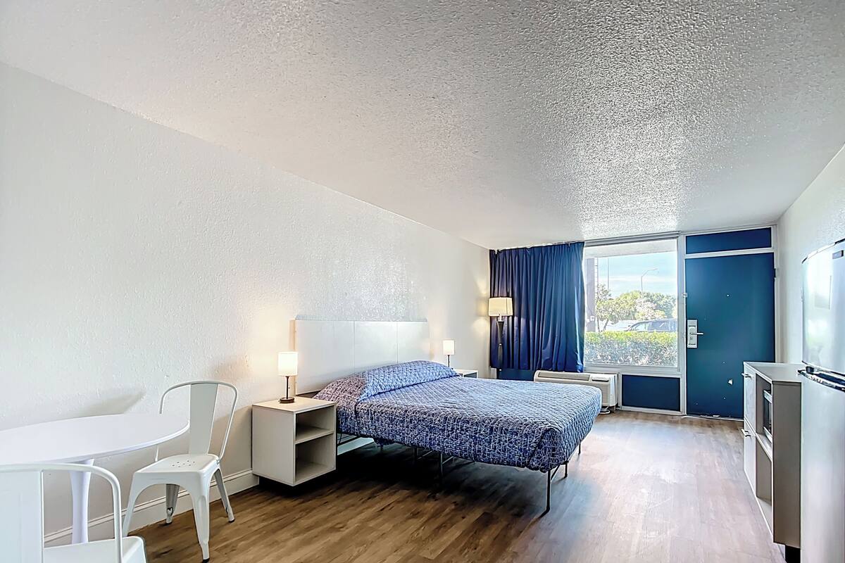 Ein Hotelzimmer mit Bett, Schreibtisch, Stuhl, kleinem Tisch, Lampe und einem Fenster mit Vorhängen.