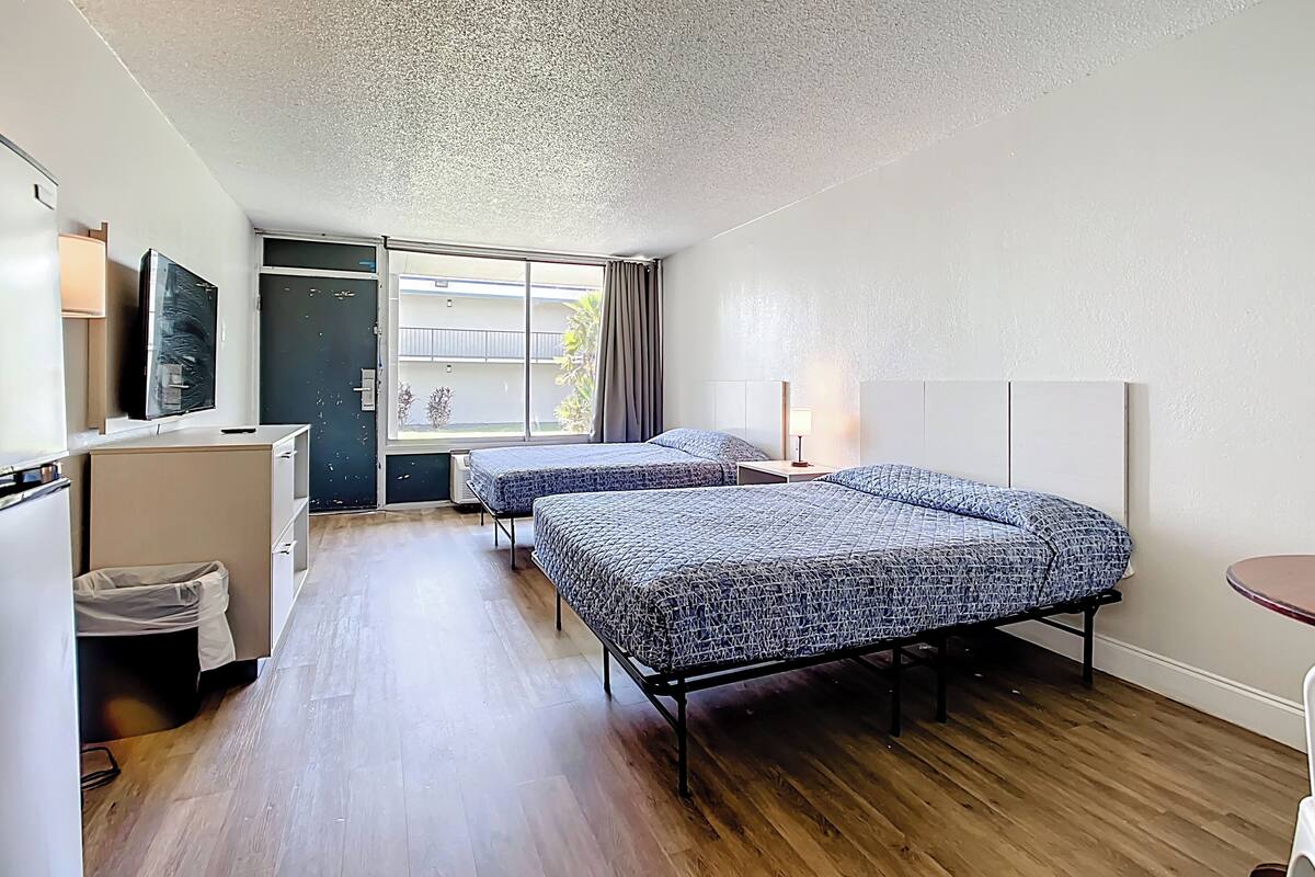Ein Hotelzimmer mit zwei Betten, einem Schreibtisch, einem Fernseher und einem Fenster mit Blick ins Freie.