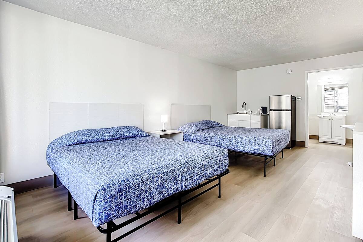 Ein Hotelzimmer mit zwei Betten, einem kleinen Nachttisch mit Lampe, einem Kühlschrank, einem Waschbecken und einem Spiegel.