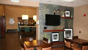 A hotel lobby with a dining area, a TV mounted on the wall, and a reception desk in the background. - Hampton Inn Phoenix/Glendale/Peoria (Peoria)