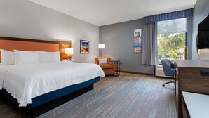 A hotel room with a large bed, a desk, and a chair. There is a window with curtains, a lamp, and a picture on the wall.