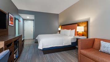 A hotel room with a bed, a desk, a chair, and a nightstand with a lamp.