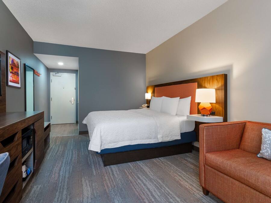 A hotel room with a bed, a desk, a chair, and a nightstand with a lamp.