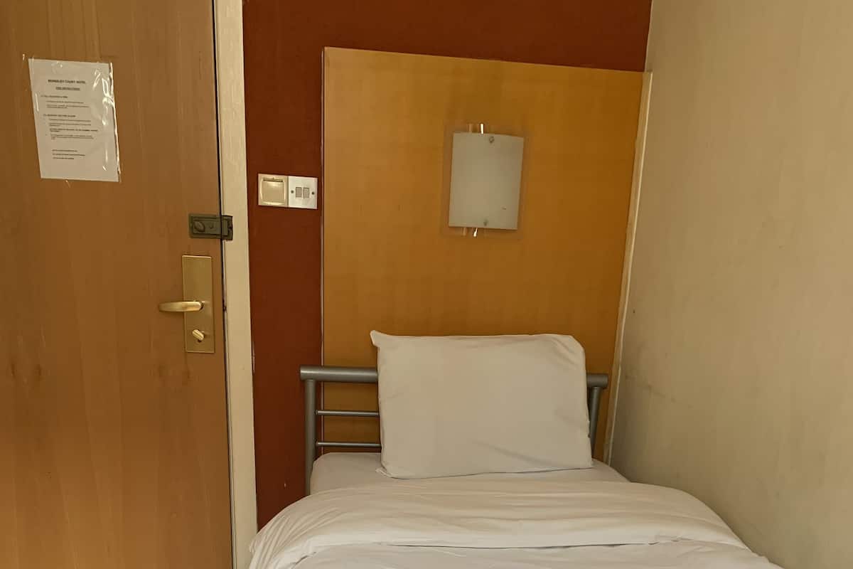 Un lit simple avec du linge de lit blanc et une serviette pliée, une porte en bois avec une pancarte, et un luminaire fixé au mur.