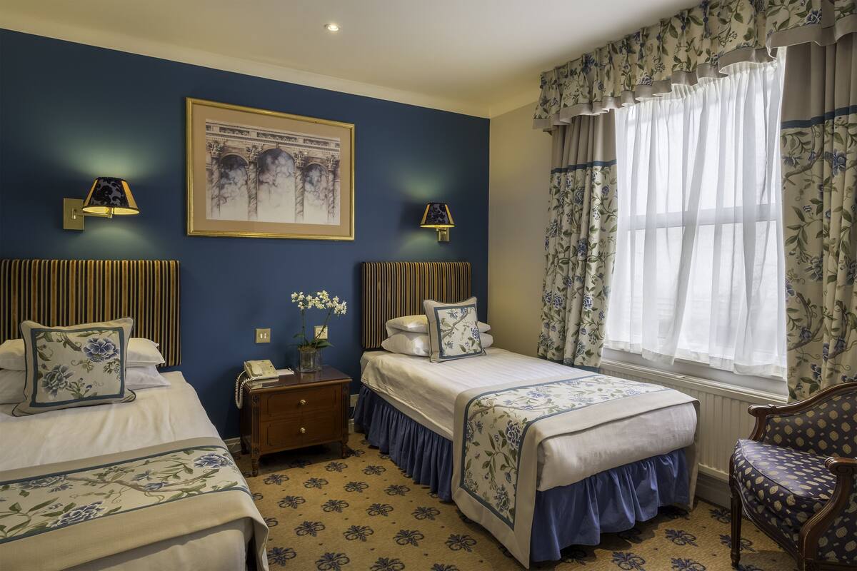 A hotel room with two beds, a nightstand, a chair, and a window with floral curtains.