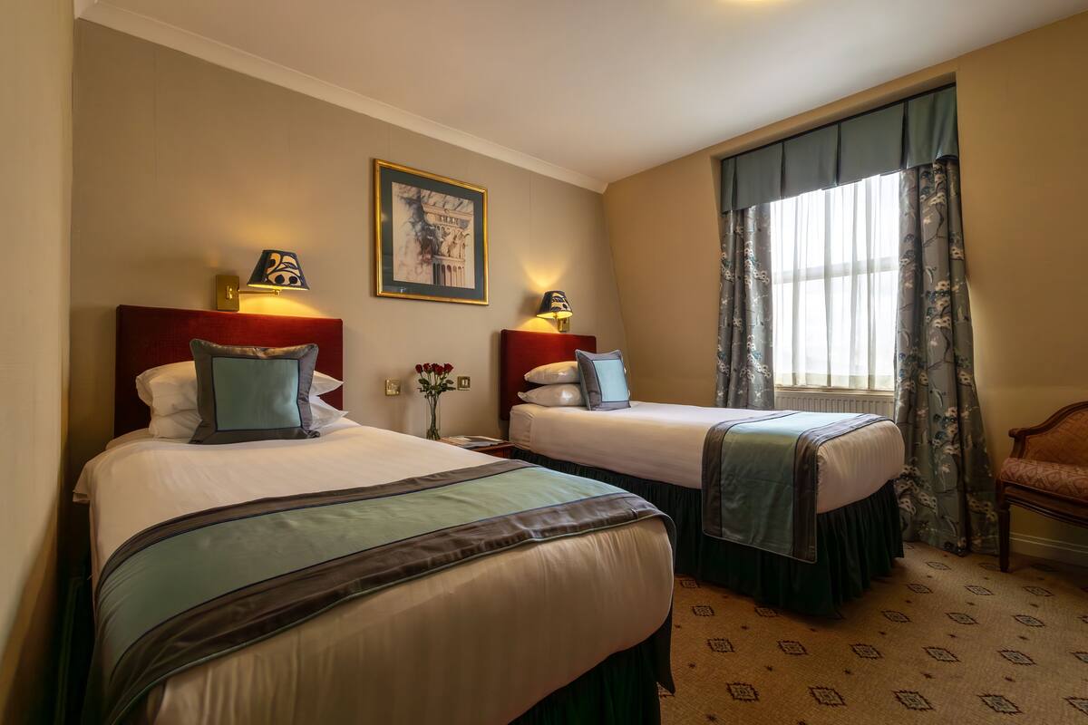 A hotel room with two beds, a nightstand with a lamp, a framed picture on the wall, and a window with curtains.