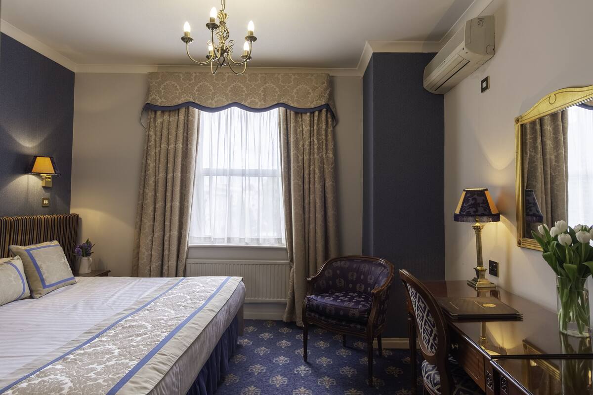 A hotel room with a bed, a chair, a desk, a lamp, a mirror, and a window with curtains.