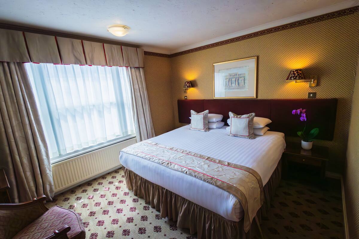 A hotel room with a large bed, a window with curtains, a radiator, a bedside table with a lamp, and a framed picture on the wall.