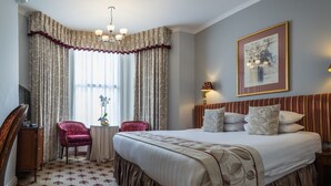 A hotel room with a large bed, a desk with a chair, a small table with a vase of flowers, and a painting on the wall. - London Lodge Hotel (London)