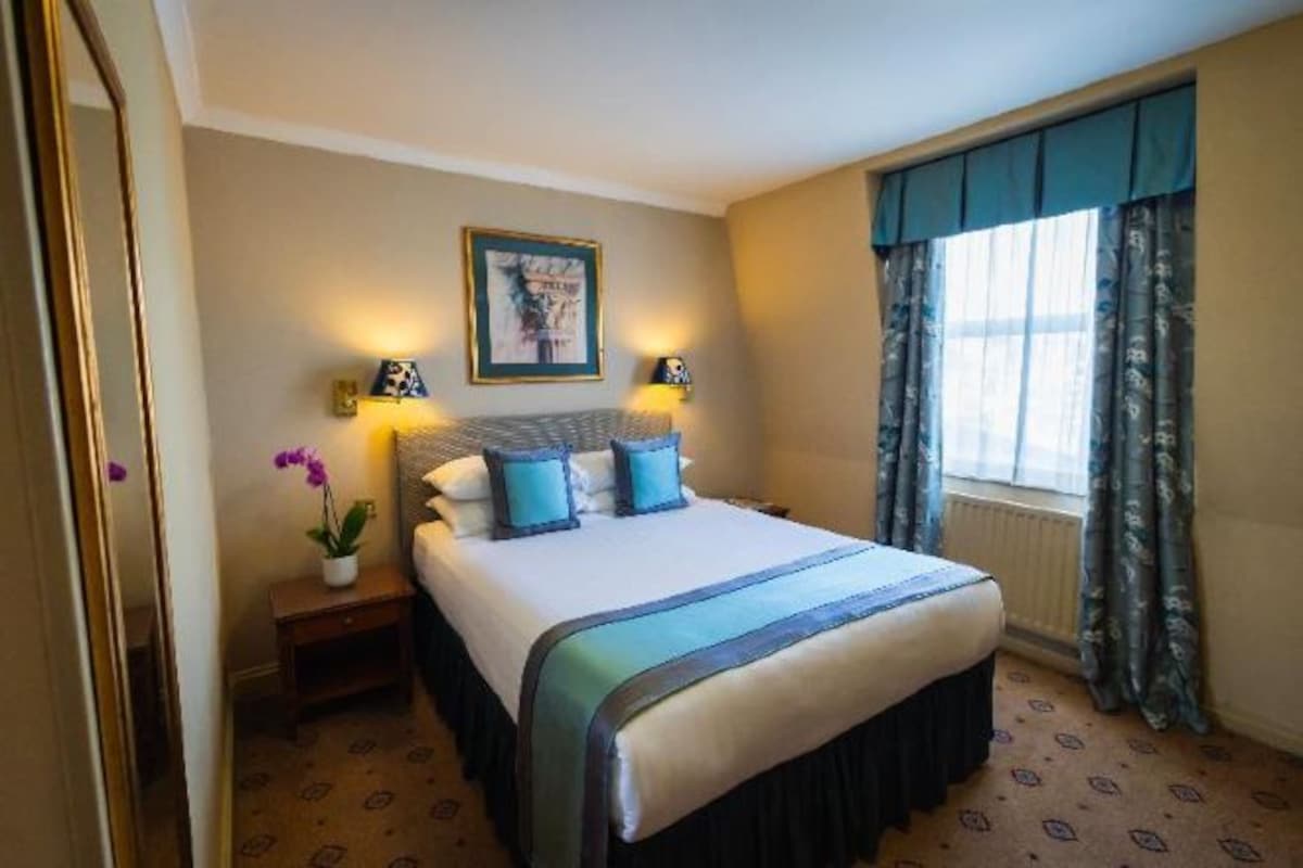 Een hotelkamer met een bed, bedlampjes, een nachtkastje, een raam met gordijnen en een ingelijste foto aan de muur.