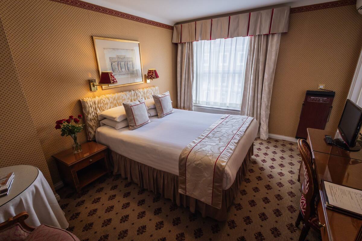 A hotel room with a large bed, a bedside table with a lamp, a small table with a vase of flowers, and a desk with a computer and papers.
