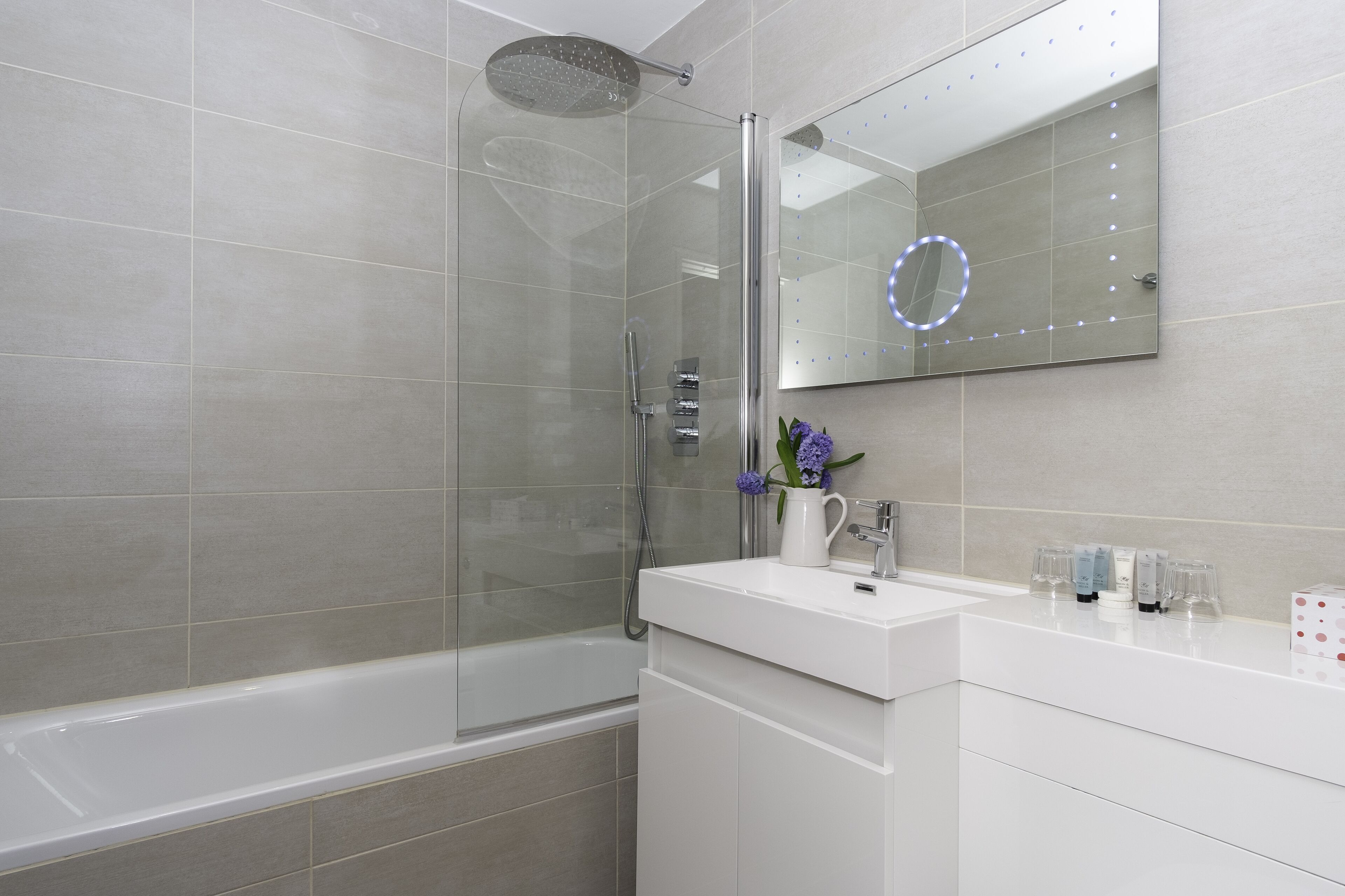 A modern bathroom with a glass-enclosed shower, a white vanity with a mirror, and a vase of purple flowers.