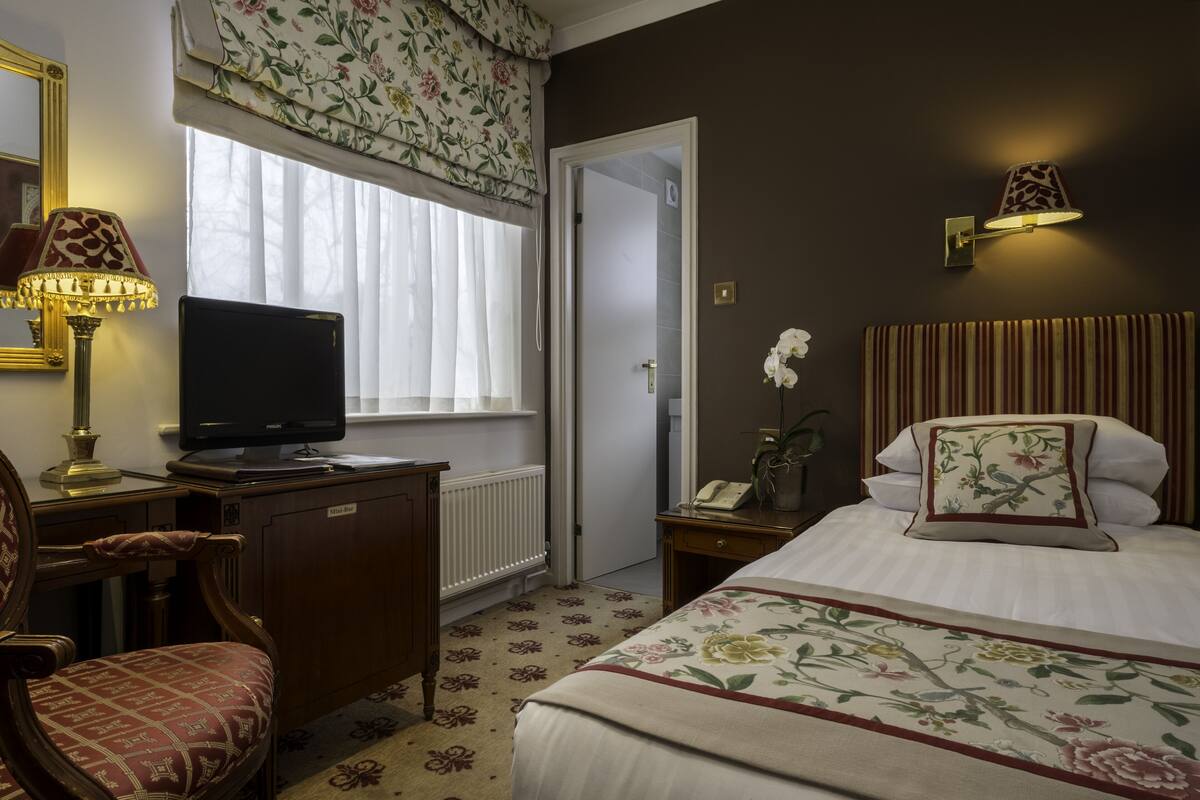 A hotel room with a bed, a desk with a television, a chair, a bedside table with a lamp, and a window with curtains.