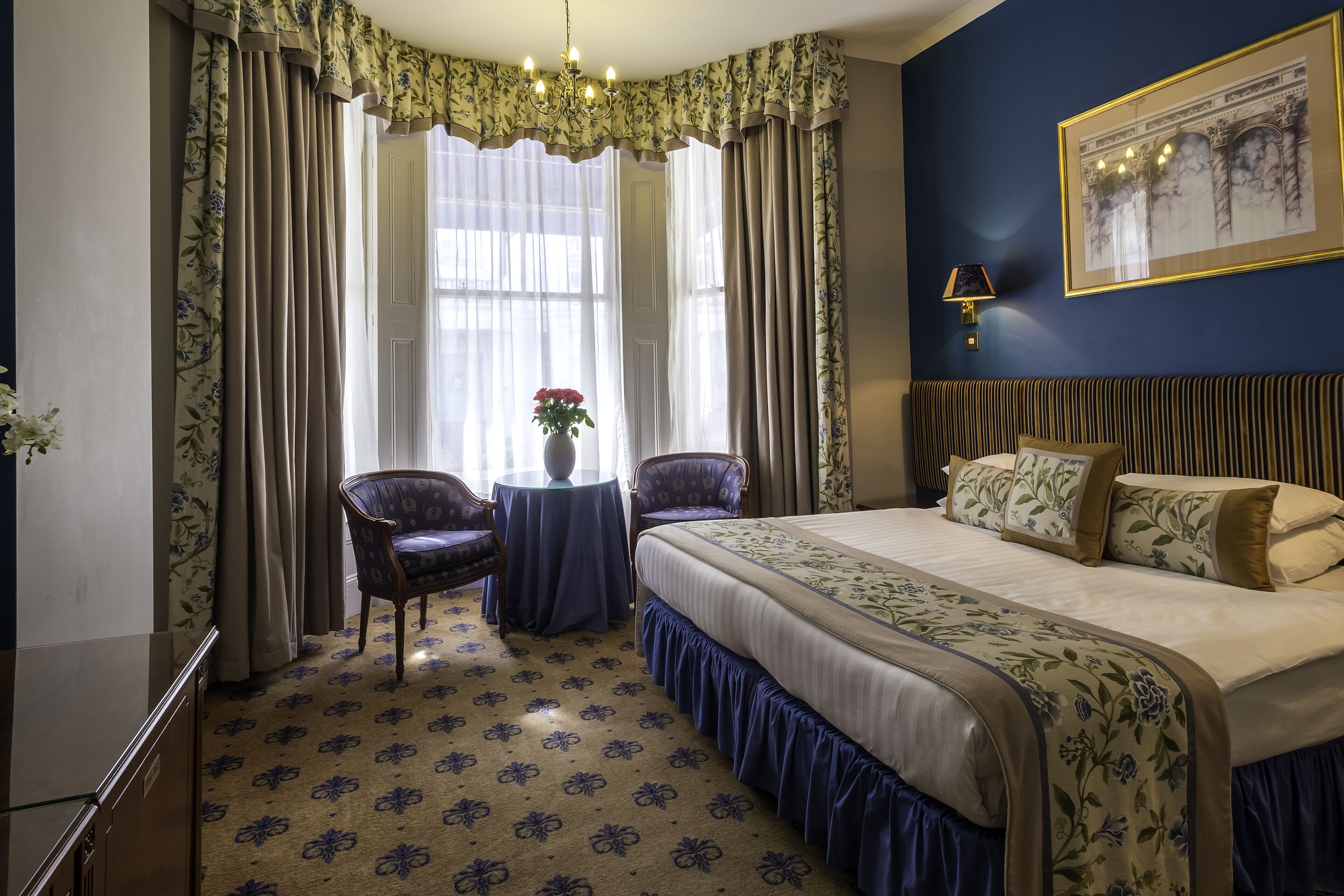 A bedroom with a large bed, two chairs, a small table with flowers, and a chandelier.
