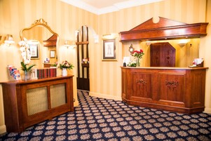 Eine Hotelhalle mit gemustertem Teppich, einem Holzbüro, einem Spiegel und gerahmten Bildern an der Wand.