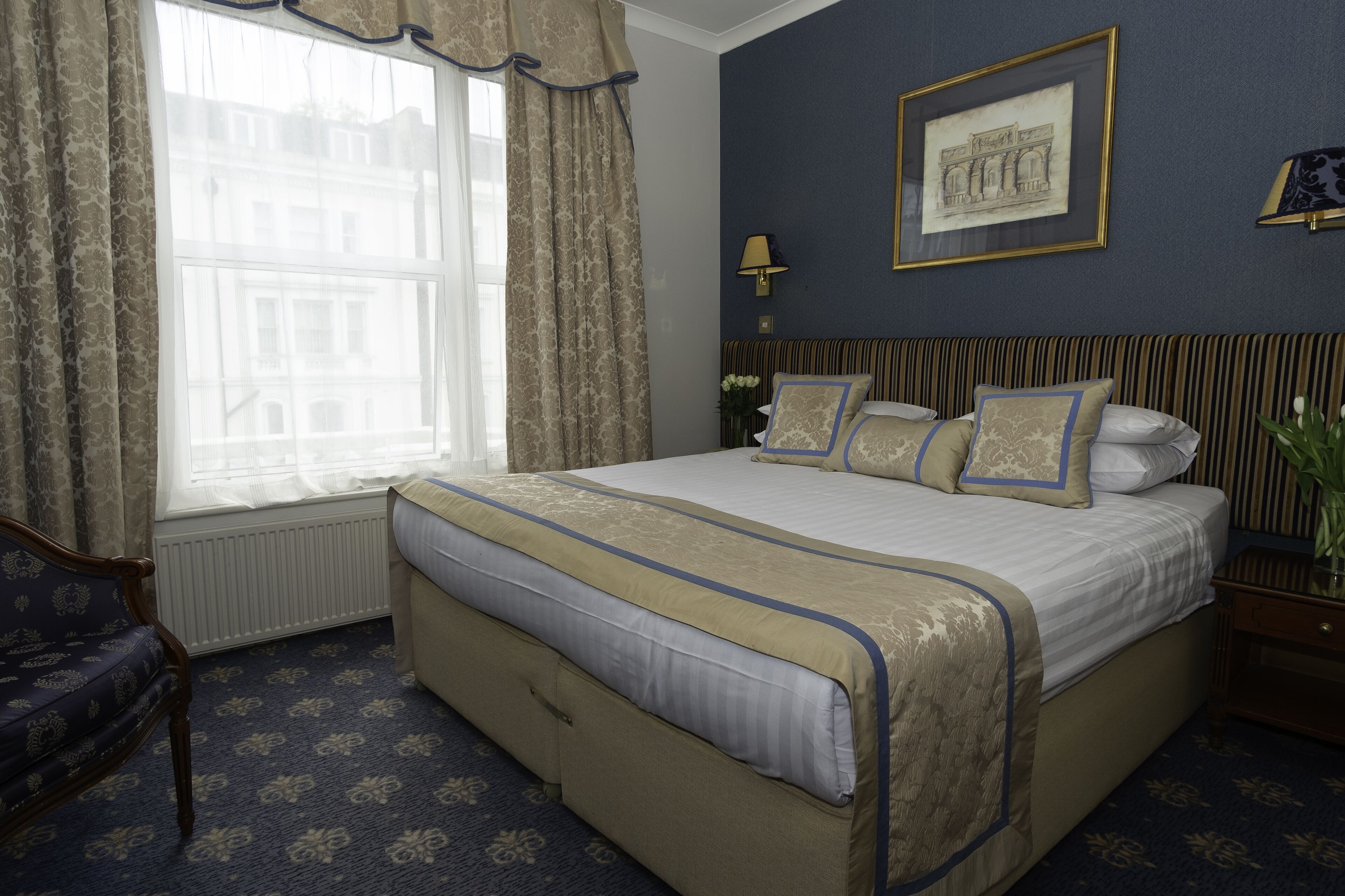 A hotel room with a large bed, a window with curtains, a framed picture on the wall, and a nightstand with a lamp.
