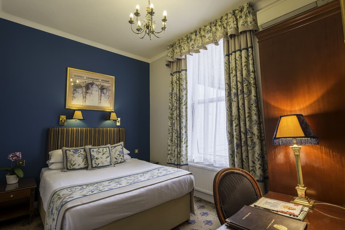 A hotel room with a bed, a desk with a lamp, a chair, a window with curtains, and a chandelier.