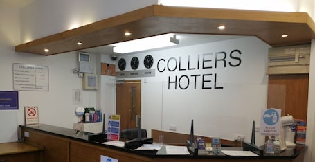 A hotel reception desk with a sign reading 'Colliers Hotel,' clocks displaying Tokyo, New York, and London times, a no-smoking sign, and a hand sanitizer station.