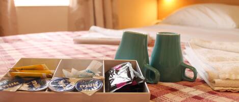 A neatly made bed with a checkered bedspread, a green mug, and a box of toiletries on a hotel room table.