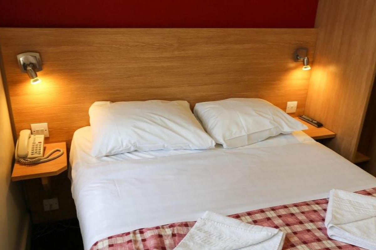 Hotellihuone, jossa on puinen sängynpääty, kaksi yöpöytävalaisinta, puhelin ja ruutukuviollinen päiväpeite.